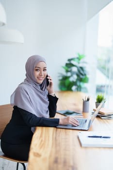 Beautiful Muslim woman talking on the phone and using a computer on her desk.