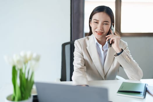 Portrait of a young Asian woman showing a smiling face as she uses her phone, computer and financial documents on her desk in the early morning hours.