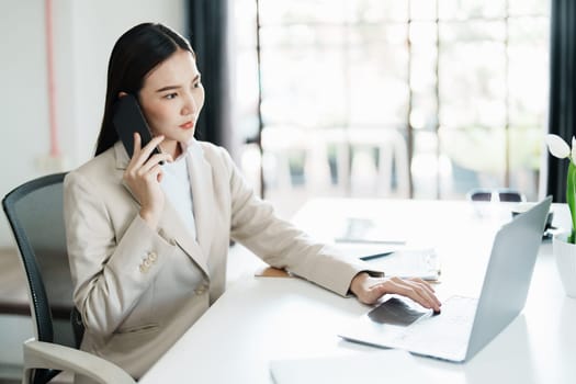 Portrait of a young Asian woman showing a smiling face as she uses her phone, computer and financial documents on her desk in the early morning hours.