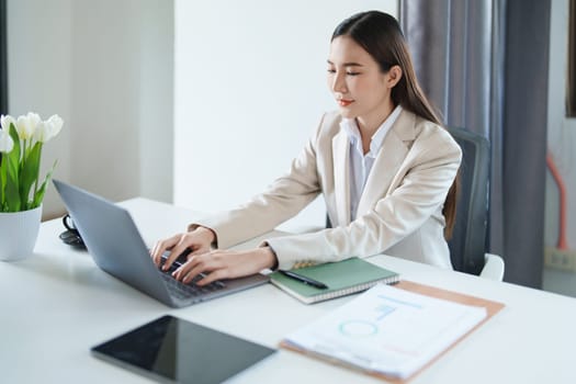 Portrait of a young Asian woman showing a smiling face as she using computer and financial documents on her desk in the early morning hours.