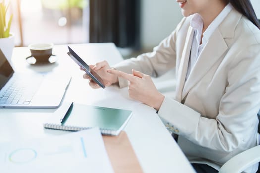 Portrait of a young Asian woman showing a smiling face as she uses her phone, computer and financial documents on her desk in the early morning hours.