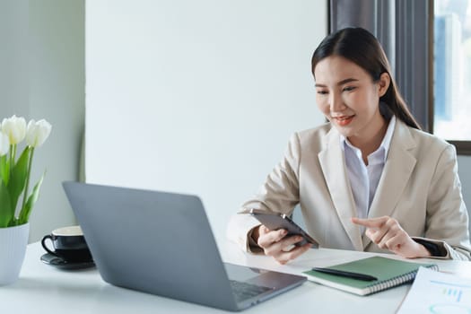 Portrait of a young Asian woman showing a smiling face as she uses her phone, computer and financial documents on her desk in the early morning hours.