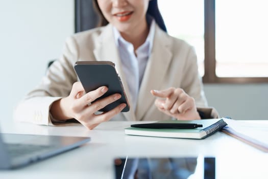 Portrait of a young Asian woman showing a smiling face as she uses her phone, computer and financial documents on her desk in the early morning hours.