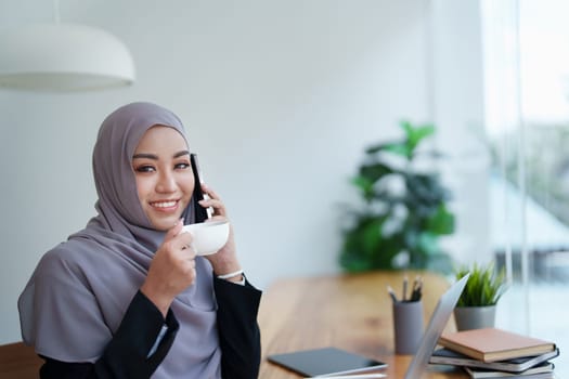 Beautiful Muslim woman talking on the phone and using computer on top while having coffee while working.