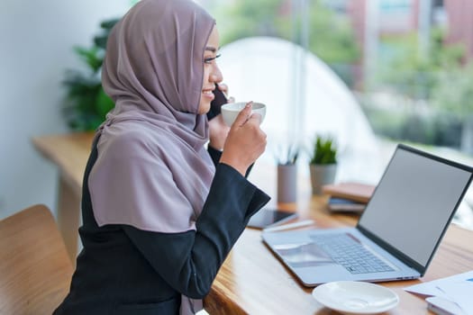 Beautiful Muslim woman talking on the phone and using computer on top while having coffee while working.
