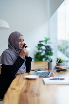 Beautiful Muslim woman talking on the phone and using computer on top while having coffee while working.