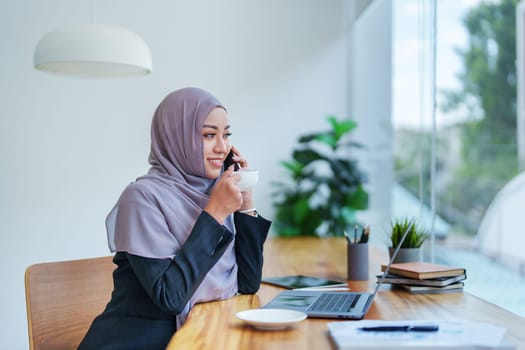 Beautiful Muslim woman talking on the phone and using computer on top while having coffee while working.