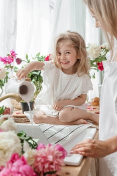 A little blonde girl with her mom on a kitchen countertop decorated with peonies. The concept of the relationship between mother and daughter. Spring atmosphere.