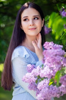 A beautiful girl with long hair stands with lilac flowers, in the spring in the park, on a sunny day. Vertical. Close up