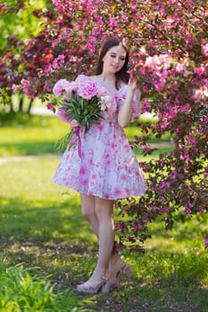 Beautiful girl, stands with a large bouquet of peonies, near pink flowering trees in the garden, in spring on a sunny day. Copy space. Vertical