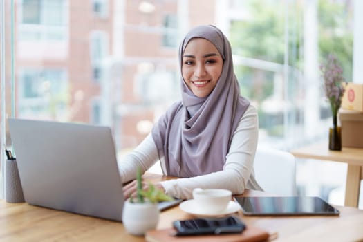 A beautiful Muslim woman with a smiling face in the morning drinking coffee and using a computer.