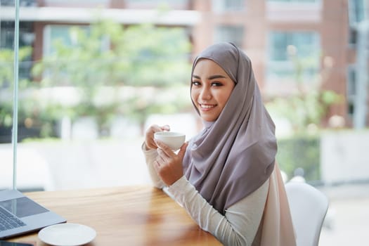 A beautiful Muslim woman with a smiling face in the morning drinking coffee and using a computer.