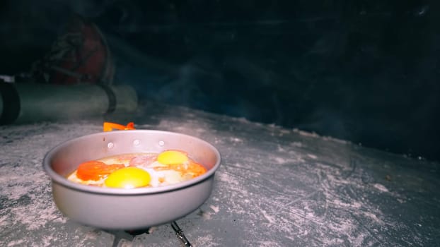 A guy cooks food on gas inside an ice cave. The climber lights the burner, cuts sausage and tomato. Breaks the eggs and fry in a frying pan. Steam from food. The ice wall is blue and the floor is gray