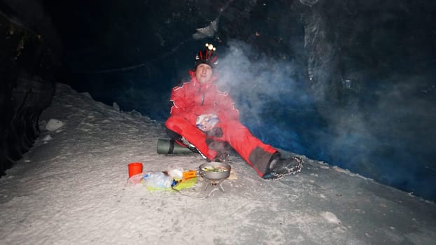 A guy cooks food on gas inside an ice cave. The climber lights the burner, cuts sausage and tomato. Breaks the eggs and fry in a frying pan. Steam from food. The ice wall is blue and the floor is gray