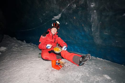 A mountaineer caver eats in a deep ice cave. Pours hot tea from a thermos, steam is coming. Eats from a frying pan, enjoys the food. Behind is a huge wall of ice with a dark blue shade. In mountains
