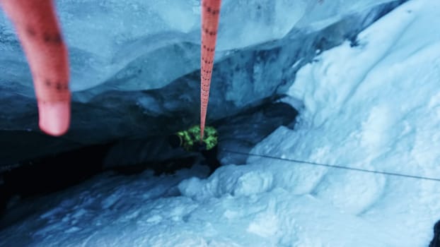 A climber descends by a safety rope into an ice cave in a glacier. The gray-blue color of ice. White snow. The guy has a bright jacket. Red rope. A dangerous descent into the unknown.