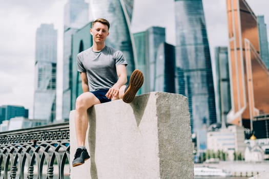 Tired Young man runner sitting on stairs and relaxing after sport training. Holding water bottle while doing fitness workout in summer city urban street, cloudy sky