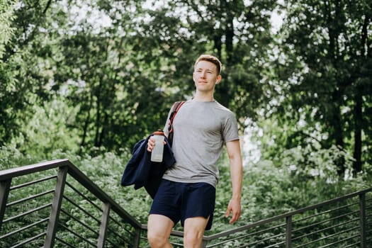 Tired Young man runner in headphones and backpack walk and relaxing after sport training. Holding water bottle while doing fitness workout in summer sunny green park.