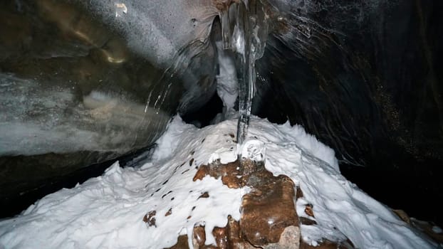 The black transparent color of ice in the cave. Small stones hang down in places. There are frozen air bubbles and large rocks in the ice. There is white snow. Icicles hang down. Small grains of snow