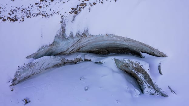 A huge ice wall from a glacier in the mountains. Everything is covered with snow and large stones. Top view from the drone on the cracks in the ice. The glacier breaks, the ice flakes off. Almaty