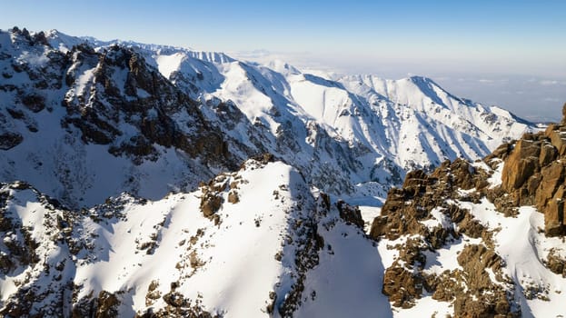 High snow-capped mountains among glaciers. Aerial view from a drone on a rocky gorge. The glacier is covered with snow and rocks. The sky is blue and the sun is shining brightly. The ice is cracking