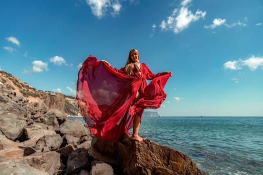 Red dress sea woman. A blonde with flowing hair in a long flowing red dress stands on a rock near the sea. Travel concept, photo session at sea.
