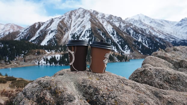 Paper cups with coffee on the background of a mountain lake. The color of the water is blue. A green forest grows on the hills. The high peaks are covered with snow. The cups are on a stone. Romance