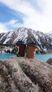 Paper cups with coffee on the background of a mountain lake. The color of the water is blue. A green forest grows on the hills. The high peaks are covered with snow. The cups are on a stone. Romance