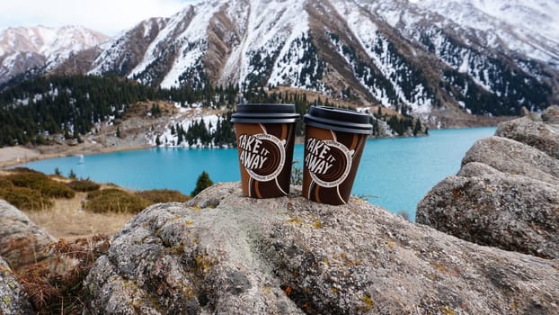 Paper cups with coffee on the background of a mountain lake. The color of the water is blue. A green forest grows on the hills. The high peaks are covered with snow. The cups are on a stone. Romance