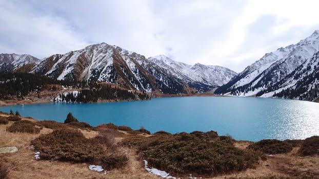 A mountain lake with blue water in winter. The sun's rays are reflected from the water. Green forest and grass grow on the hills. There are stones lying. The high peaks are covered with snow. Almaty