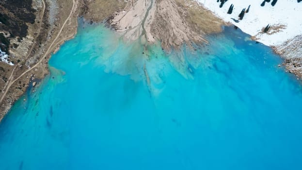 A lake in the mountains with turquoise blue water. Drone view of clear water, coniferous trees and snowy mountains. People walk along the shore, low bushes grow. Big Almaty lake. Kazakhstan