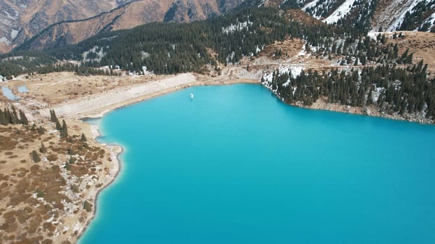 A lake in the mountains with turquoise blue water. Drone view of clear water, coniferous trees and snowy mountains. People walk along the shore, low bushes grow. Big Almaty lake. Kazakhstan
