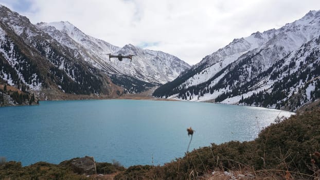 A mountain lake with blue water in winter. The sun's rays are reflected from the water. Green forest and grass grow on the hills. There are stones lying. The high peaks are covered with snow. Almaty