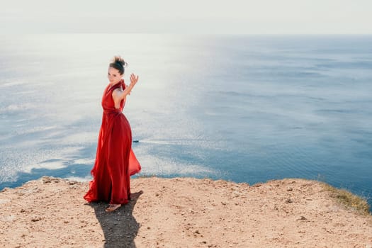 Side view a Young beautiful sensual woman in a red long dress posing on a rock high above the sea during sunrise. Girl on the nature on blue sky background. Fashion photo.