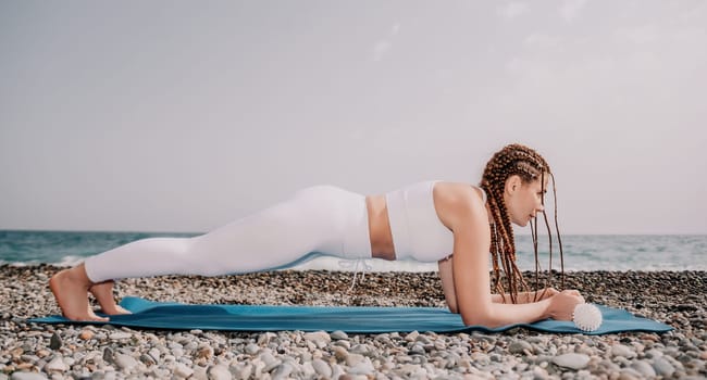 Woman yoga sea. Well looking middle aged woman with braids dreadlocks in white leggings and tops doing stretching pilates on yoga mat near sea. Female fitness yoga routine concept. Healthy lifestyle