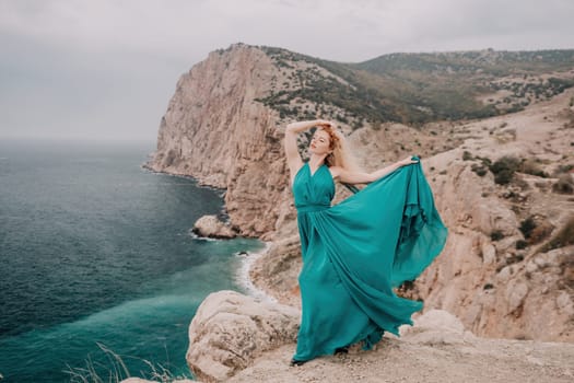 Side view a Young beautiful sensual woman in a mint long dress posing on a volcanic rock high above the sea during sunset. Girl on the nature on overcast sky background. Fashion photo