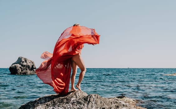 Woman travel sea. Young Happy woman in a long red dress posing on a beach near the sea on background of volcanic rocks, like in Iceland, sharing travel adventure journey