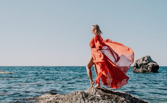 Woman travel sea. Young Happy woman in a long red dress posing on a beach near the sea on background of volcanic rocks, like in Iceland, sharing travel adventure journey