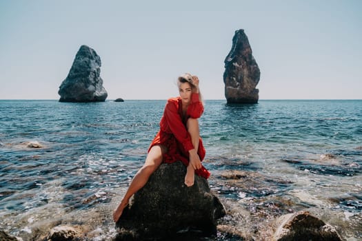 Woman travel sea. Young Happy woman in a long red dress posing on a beach near the sea on background of volcanic rocks, like in Iceland, sharing travel adventure journey