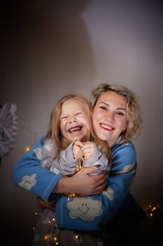 Cute mother and daughter in pajamas having fun in the room with Christmas garlands and white background. The tradition of decorating the house for holidays. Happy childhood and motherhood