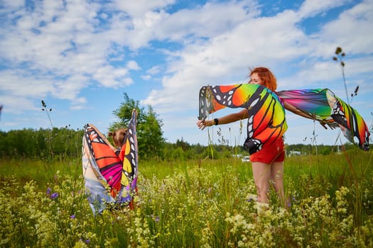 Happy female family with red haired mother and daughter with bright butterfly wings having fun on green and yellow meadow full of grass and flowers in sunny summer day. Concept family love