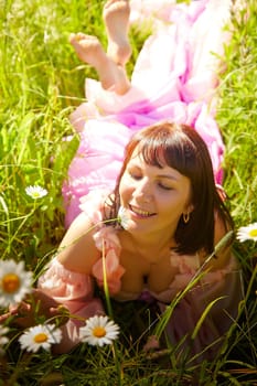 Beautiful girl in lush pink ball gown in green field during blooming of flowers and blue sky on background. Model posing on nature landscape as princess from fary tale