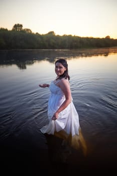 Slavic plump plump chubby girl in long white dress on the feast of Ivan Kupala with flowers and water in a river or lake on summer evening