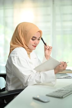 Thoughtful muslim businesswoman wearing hijab working with statistics at her office desk.