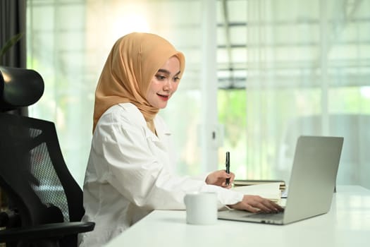 Portrait of young young muslim woman in hijab using laptop sitting at desk, writing in notebook.