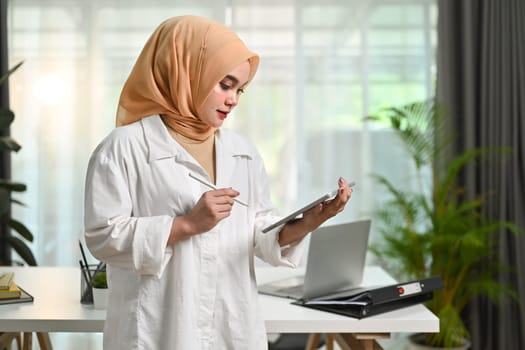 Muslim businesswoman wearing hijab using digital tablet while standing in an office boardroom.