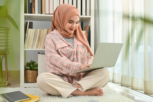 Happy joyful muslim woman wearing hijab using laptop for distant studying, online communication and remote work.