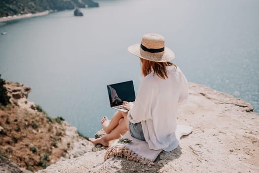 Successful business woman in yellow hat working on laptop by the sea. Pretty lady typing on computer at summer day outdoors. Freelance, travel and holidays concept.