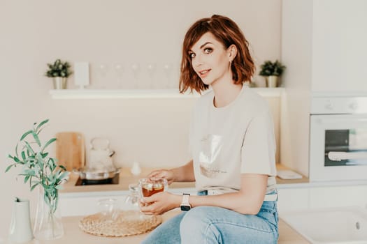 Woman kitchen tea. Carefree woman rests in a cozy kitchen, drinks tea, sits on the table and dreams, free space. Woman holding cup while enjoying hot drink at home.