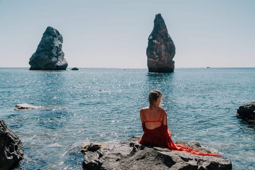 Woman travel sea. Young Happy woman in a long red dress posing on a beach near the sea on background of volcanic rocks, like in Iceland, sharing travel adventure journey